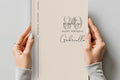 A person holds a cream-colored 80th birthday letter or notebook with elegant black script and serif text reading 'eighty happy birthday Gabriella' against a neutral gray background.