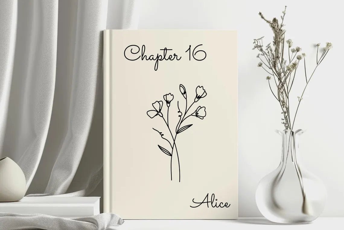 A cream-colored hardcover book standing upright on a tabletop, printed with the text 'Chapter 16' and 'Alice' in black script, with a simple black floral line drawing in the center. The book is styled in a bright, minimalist room setting with draped white fabric, a small white vase, and dried stems in a clear glass vase.