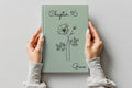 A person holds a sage green hardcover journal with 'Chapter 16' and the name 'Grace' printed on the cover, featuring a minimalist black line drawing of a flower. The person is wearing a light gray long-sleeve top, and the background is a clean, neutral gray wall.