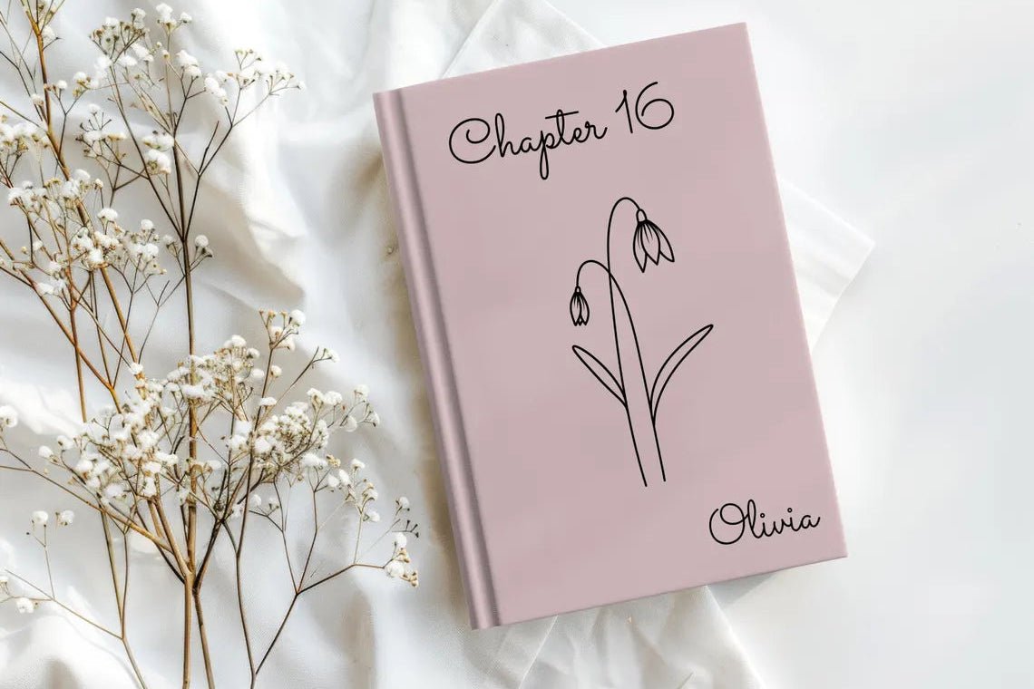 A pink hardcover journal or notebook with 'Chapter 16' and a simple flower line drawing on the cover, styled on white draped fabric with baby's breath flowers beside it. The name 'Olivia' is printed in script near the bottom.
