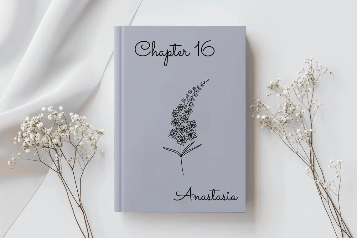 A personalized hardcover journal with a light gray cover, featuring the text 'Chapter 16' and 'Anastasia' with a black botanical illustration in the center. The journal is styled in a flat-lay arrangement with white baby's breath flowers on both sides against a white background with soft draped fabric.