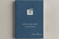 A hardcover movie review logbook in slate blue with a white clapperboard icon and white text, including the name James Brown, shown on a neutral light gray background.