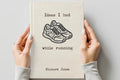 A person holds a cream-colored hardcover notebook with a black typewriter-style illustration of running shoes and the text 'Ideas I had while running' and 'Richard Jones' on the cover, against a light gray background.