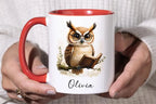 A person holding a white ceramic mug with a red handle and red interior. The mug features a printed illustration of a brown and beige owl reading a book, with the name 'Olivia' written below in black script.
