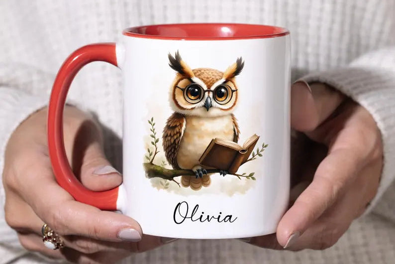 A person holding a white ceramic mug with a red handle and red interior. The mug features a printed illustration of a brown and beige owl reading a book, with the name 'Olivia' written below in black script.